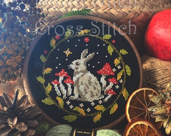 May include: A cross stitch embroidery hoop with a black background featuring a white rabbit surrounded by green leaves and red mushrooms. The hoop is surrounded by yarn, dried fruit, and a pine cone.