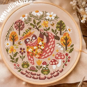 May include: Cross-stitch embroidery in a wooden hoop with a sloth design, surrounded by flowers, trees, and mushrooms. The design is on a beige fabric. The text "Mama Witch x Stitch" is visible.