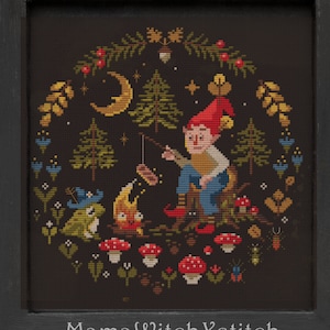 May include: A cross-stitch pattern featuring a gnome sitting by a campfire with a frog and a firefly. The scene is surrounded by a wreath of forest foliage, mushrooms, and other woodland elements. The text "MamaWitchXStitch" is at the bottom of the image.