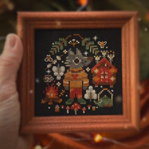 May include: A framed cross-stitch piece on black fabric featuring a central wolf character in a yellow sweater, surrounded by a campfire, a small house, mushrooms, and pine branches. The text MAMA WITCH XSTITCH appears at the bottom of the image.