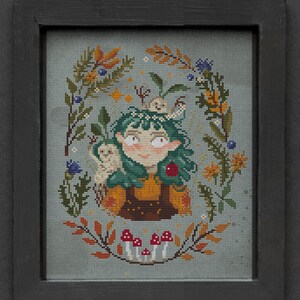 Puede incluir: Un patrón de punto de cruz con una niña de pelo verde con una corona de flores, rodeada de criaturas del bosque y follaje. El patrón está enmarcado en un marco negro y el texto "Mama Witch" es visible debajo del marco.