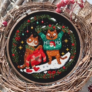 May include: A cross-stitch embroidery hoop with a black background featuring two cats wearing Christmas jumpers. The cats are surrounded by a wreath of green pine branches with red berries and baubles. The text "Ho Ho" is on one of the jumpers.