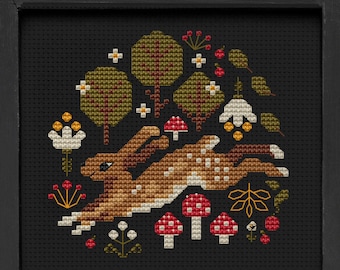 May include: Cross stitch pattern featuring a brown rabbit running through a forest with mushrooms, flowers, and trees. The pattern is framed in a black frame and the text "Mama Witch Xstitch" is below the frame.