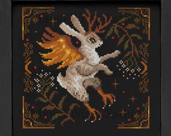 May include: A cross-stitch pattern featuring a white rabbit with antlers and golden wings, surrounded by a black background with gold and green floral details.