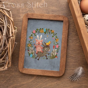 May include: A cross stitch pattern depicting a pink pig riding a donkey, encircled by a wreath of flowers and bees. The pattern is framed in a dark wood frame and presented on a wooden surface.
