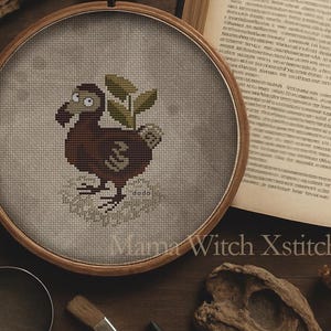 Op de afbeelding: Een bruine en witte kruissteek-borduurwerk van een dodo met groene bladeren in een ronde houten borduurring. De ring ligt op een houten tafel met een boek, een vergrootglas en andere decoratieve items.