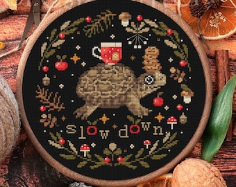 Op de afbeelding: Een zwarte borduurring met een kruissteekontwerp van een schildpad die een kopje warme chocolademelk en een stapel koekjes draagt. Het ontwerp is omringd door een krans van bladeren, bessen en paddenstoelen. De tekst "slow down" is onder de schildpad geborduurd.
