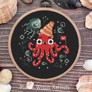 Puede incluir: Un aro de bordado de punto de cruz con fondo negro que presenta un pulpo rojo con un sombrero marrón y naranja. El pulpo tiene grandes ojos blancos y está rodeado de diseños florales verdes y blancos.