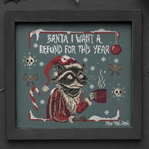 May include: Cross-stitch artwork in a black frame featuring a raccoon dressed as Santa Claus, holding a mug. The text reads "SANTA I WANT A REFUND FOR THIS YEAR." The design includes snowflakes, skulls, and a candy cane.