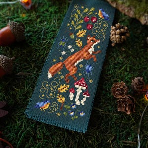 Può includere: Segnalibro a punto croce con una volpe che salta, funghi e un uccellino blu su sfondo verde scuro. Il disegno include elementi floreali e insetti. Il testo "MAMA WITCH XSTITCH" è visibile.