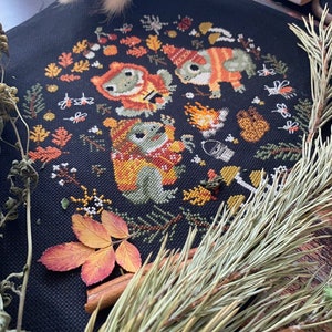 May include: Cross-stitch pattern featuring three frogs wearing hats and scarves around a campfire with autumn leaves and other woodland elements.