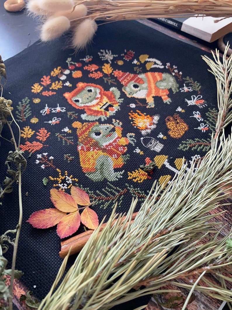 May include: Cross-stitch pattern featuring three frogs wearing hats and scarves around a campfire with autumn leaves and other woodland elements.