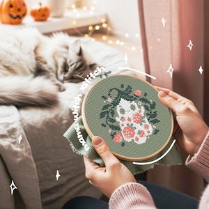 May include: A cross-stitch embroidery hoop featuring a Halloween-themed design. The design includes a smiling ghost with floral accents in pink and green. The text "mama witch xstitch" is visible on the fabric. The background includes a sleeping cat and decorative pumpkins.