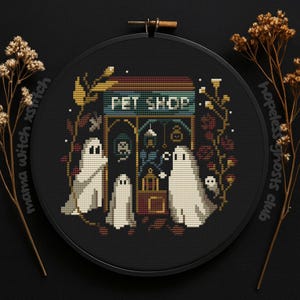 Op de afbeelding: Een kruissteekborduurwerk met een "Pet Shop" scène met geesten. Het ontwerp omvat een winkelgevel met hangende lantaarns en een bord. De geesten zijn wit en de achtergrond is zwart. De borduursel is ingelijst in een zwarte hoepel.