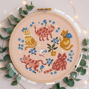 May include: Cross-stitch pattern featuring five stylized cats in various poses, surrounded by blue flowers and a small potted plant. The design is set against a light pink background within a wooden embroidery hoop. Text reads "mama witch xstitch" and "CROSS STITCH PATTERN".