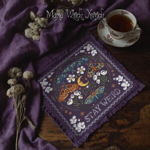 May include: A purple cross-stitch featuring a circular design with moths, flowers, and a crescent moon. The text "STAY WEIRD" is stitched in white. The image is on a purple cloth with a cup of tea in the background.