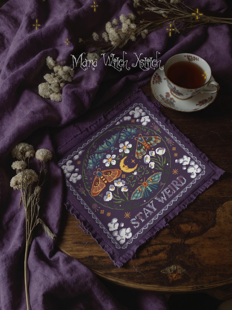 May include: A purple cross-stitch featuring a circular design with moths, flowers, and a crescent moon. The text "STAY WEIRD" is stitched in white. The image is on a purple cloth with a cup of tea in the background.