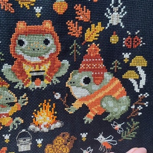 May include: Cross-stitch embroidery featuring two frogs wearing orange and green sweaters, sitting by a campfire with a bucket and logs. The background is black with autumn leaves, mushrooms, and flying insects.