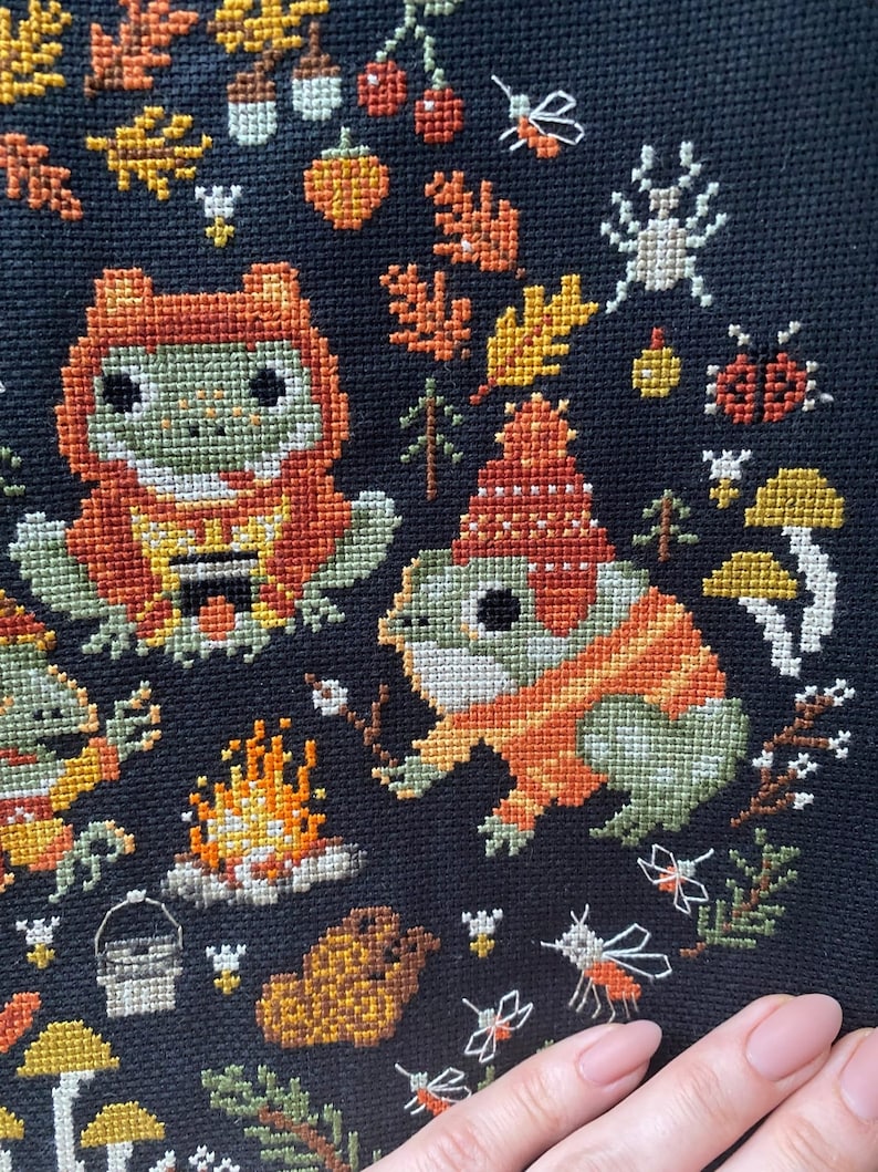 May include: Cross-stitch embroidery featuring two frogs wearing orange and green sweaters, sitting by a campfire with a bucket and logs. The background is black with autumn leaves, mushrooms, and flying insects.