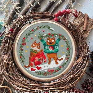 May include: Cross-stitch embroidery hoop with a winter scene of two cats wearing Christmas sweaters. The cats are standing in the snow with a green wreath above them. The text "HO HO" is on one of the sweaters.