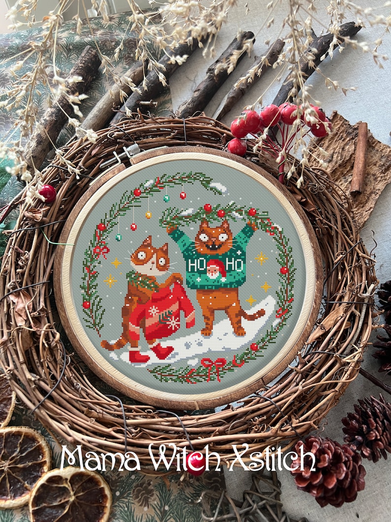May include: Cross-stitch embroidery hoop with a winter scene of two cats wearing Christmas sweaters. The cats are standing in the snow with a green wreath above them. The text "HO HO" is on one of the sweaters.