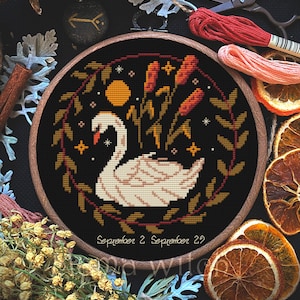 May include: A cross-stitch embroidery in a wooden hoop features a white swan, a yellow moon, and orange cattails against a black background. The text "September 2 September 29" is below the design. The design is surrounded by a brown and green wreath.