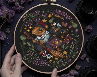 May include: A cross-stitch pattern featuring a cute squirrel dressed as a witch, flying on a broomstick with a raven and potion bottles. The design is surrounded by flowers and stars on a black background.