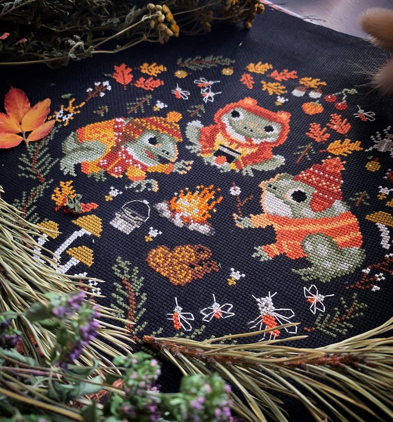 May include: Cross-stitch embroidery of three frogs wearing sweaters and hats, gathered around a campfire with a bucket and a heart. The background is black with green and orange foliage, and white fireflies.