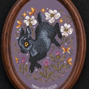 May include: An oval wooden frame containing a cross-stitch embroidery on a purple background. The design features a black rabbit with yellow eyes, surrounded by white flowers, purple thistles, and orange butterflies. Text reads MAMA WITCH XSTITCH.
