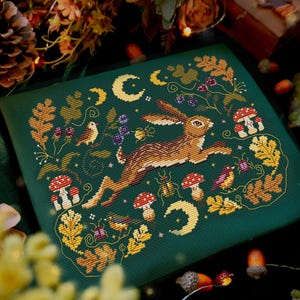 Op de afbeelding: Kruissteekontwerp op groene stof met een rennende haas, paddenstoelen, vogels en halve manen. Het ontwerp is omlijst door herfstbladeren en bessen. De tekst "MAMA WITCH XSTITCH" staat rechtsonder.