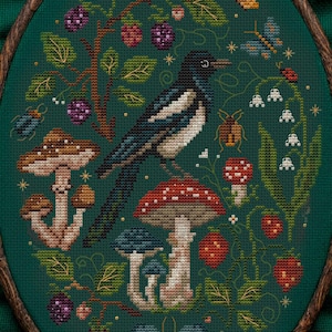 Könnte beinhalten: Kreuzstich-Stickerei mit einer Elster auf einem Pilz, umgeben von Beeren, Pilzen und Insekten. Das Design befindet sich auf einem dunkelgrünen Hintergrund in einem ovalen Holzrahmen. Der Text "MAMA WITCH XSTITCH" ist sichtbar.