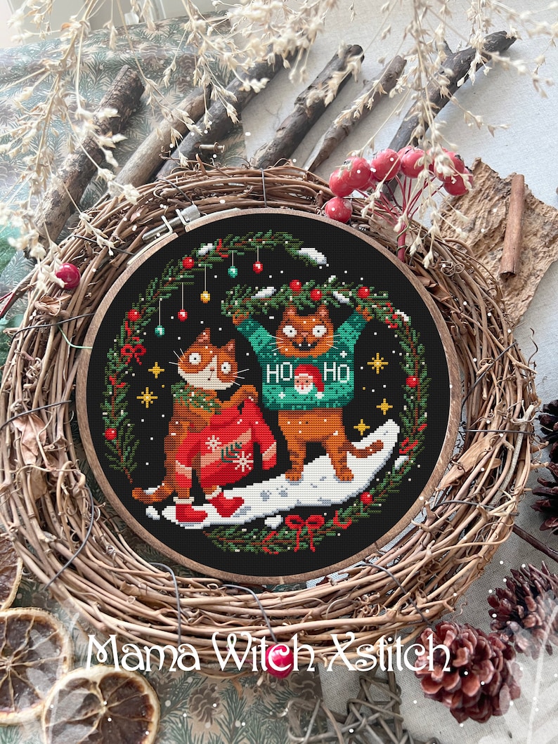 May include: A cross-stitch embroidery hoop with a black background featuring two cats wearing Christmas sweaters. The cats are surrounded by a wreath of green pine branches with red berries and ornaments. The text "Ho Ho" is on one of the sweaters.