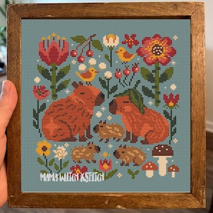 May include: Cross-stitch design showing a capybara family with flowers, birds, and mushrooms on a teal background, framed in wood. The text "Mama Witch x Stitch" is visible. The artwork is in a square format.