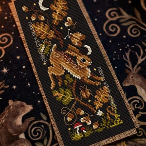 Op de afbeelding: Kruissteek bladwijzer met een bruine haas die door een bosscène springt met bladeren, eikels en paddenstoelen. De bladwijzer heeft de tekst "mama witch xstitch" en een rand.