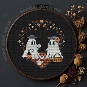 Könnte beinhalten: Eine Kreuzstichstickerei in einem Holzreifen zeigt zwei weiße Geisterfiguren mit Hüten, die Tee trinken. Das Design umfasst eine braun-orangefarbene Picknickdecke, Körbe und den Text "hopeless ghosts club".