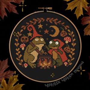 May include: Cross-stitch embroidery in a wooden hoop featuring two frogs around a cauldron. One frog wears a witch hat, the other a red cape. The design includes pumpkins, mushrooms, stars, and a crescent moon, all in autumnal colors.