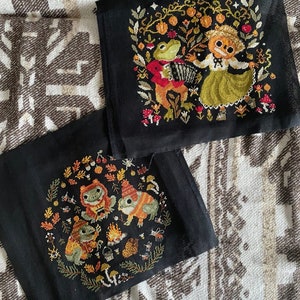 May include: Two black fabric panels with embroidered images. The top panel features a frog playing an accordion and a girl in a green dress. The bottom panel features three frogs around a campfire.