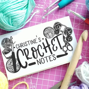 May include: A white notebook with the text "CHRISTINE'S CROCHET NOTES" in black lettering. The notebook is surrounded by colourful yarn balls, crochet hooks, and scissors on a pink cutting mat.