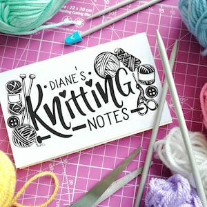 May include: A white notebook with black lettering that says "Diane's Knitting - Notes". The notebook is surrounded by balls of yarn in various colors, including pink, blue, white, and yellow. There are also knitting needles and scissors.