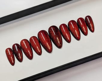 May include: Set of ten almond-shaped, press-on nails. The nails are a deep red color with a glittery, shimmering effect. They are arranged in a row inside a black rectangular box, ready for application. The nails are a beauty product.