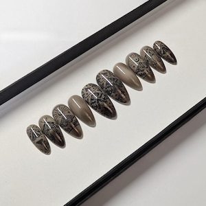 May include: A set of ten almond-shaped press-on nails in a glossy taupe finish. Each nail features a black line art design of a moth, celestial sun, moon, and stars. The nails are arranged in a row, showcasing a consistent color palette and detailed graphic illustrations on several of the individual nail pieces.