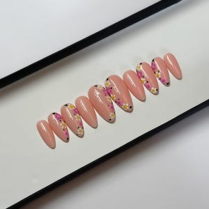 May include: A collection of almond-shaped false nails in a pale peach colour. Each nail is decorated with a floral pattern of white, yellow, and pink flowers with black detailing. The nails are presented on a white surface within a black box.