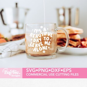 May include: A white coffee mug with the text "It's a beautiful day to leave me alone" in a white circle with small flower accents. The mug is filled with coffee and cream. The mug is on a white table with a plate of waffles and bacon in the background.