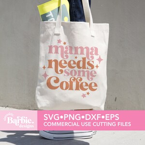 Op de afbeelding: Een witte tote bag met de tekst "mama needs some coffee" in roze en oranje letters met sterren. De tas wordt gedragen door iemand die een spijkerbroek draagt.