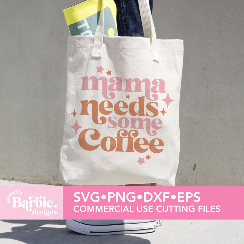 Op de afbeelding: Een witte tote bag met de tekst "mama needs some coffee" in roze en oranje letters met sterren. De tas wordt gedragen door iemand die een spijkerbroek draagt.