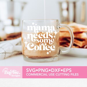 Op de afbeelding: Een heldere glazen mok met een wit ontwerp met de tekst "mama needs some coffee" met sterren rond de tekst. De mok is gevuld met koffie en room.