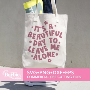 May include: A white tote bag with a pink design that says "It's a beautiful day to leave me alone." The design includes stars and flowers. The bag is being held by a person wearing jeans.