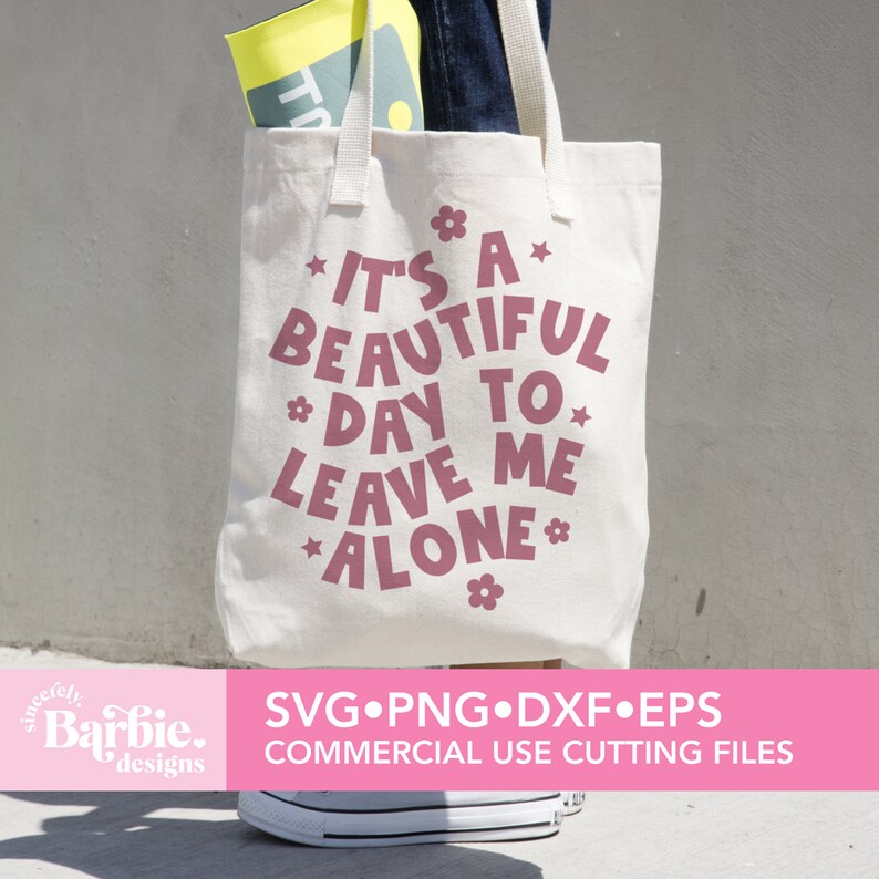 May include: A white tote bag with a pink design that says "It's a beautiful day to leave me alone." The design includes stars and flowers. The bag is being held by a person wearing jeans.