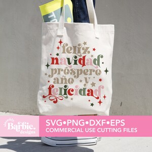 May include: A white tote bag with the text "feliz navidad prospero a&ntilde;o y felicidad" in a festive font with red, green, and gold accents. The text is surrounded by stars and swirls. The bag is being held by a person wearing blue jeans.