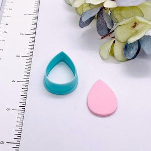 May include: A teal teardrop-shaped cookie cutter next to a pink teardrop-shaped object. The cutter is made of plastic and has a rounded base. The pink object appears to be a clay or fondant shape. A ruler is in the background.